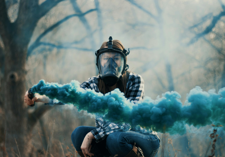 Une femme portant un masque à gaz et tenant une fusée bleue devant un fond d'arbres.