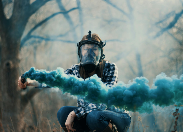 Une femme portant un masque à gaz et tenant une fusée bleue devant un fond d'arbres.