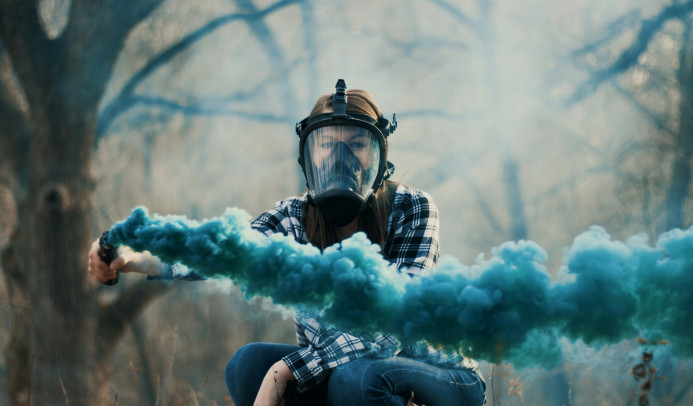 Une femme portant un masque à gaz et tenant une fusée bleue devant un fond d'arbres.