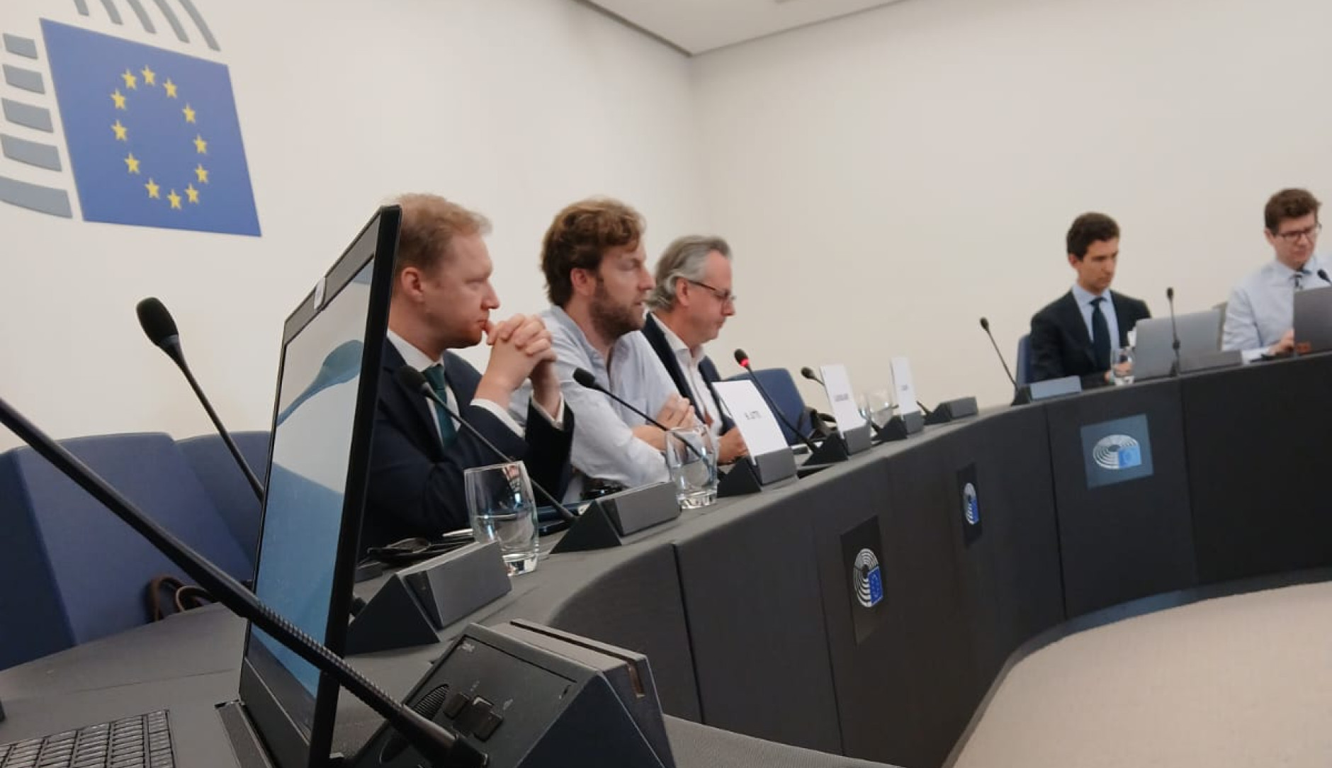 5 hommes assis derrière un bureau avec un intervenant au micro