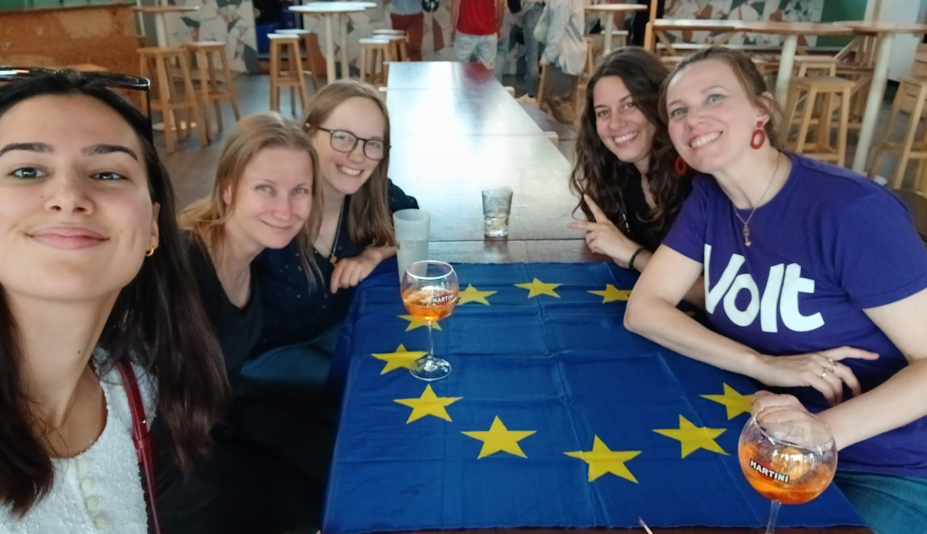 selfie 5 jeunes femmes souriantes attablées, tee shirt volt et drapeau européen
